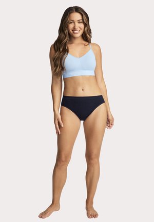 Smiling woman with long wavy hair wearing a light blue sports bra and black underwear, standing barefoot on a plain background.