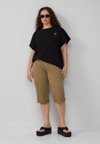 Zwarte oversized t-shirt met kleine grafische print, khaki shorts met omgeslagen mouwen, en zwarte platform sandalen. Smooth textuur, casual pasvorm, neutrale kleuren.
