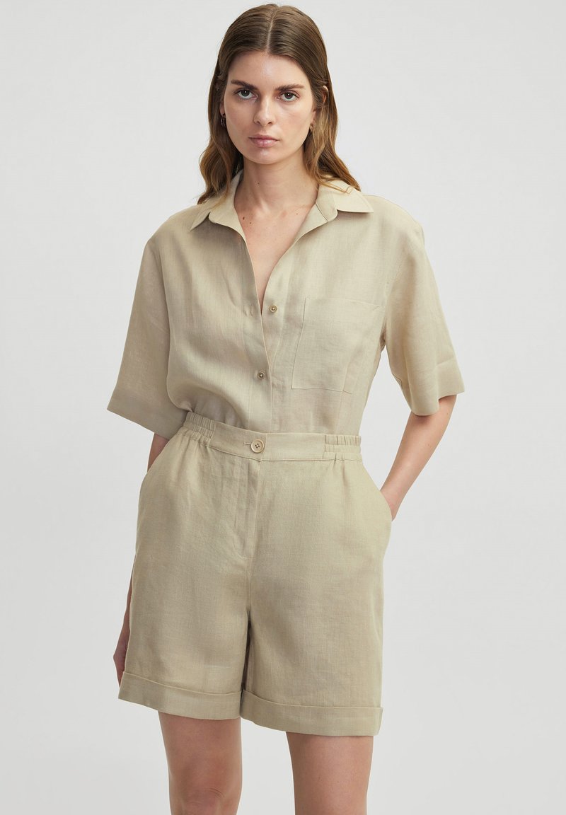 Combinaison en lin beige à manches courtes avec un col, une patte de boutonnage, une poche sur la poitrine gauche, une taille élastique et un short à revers.