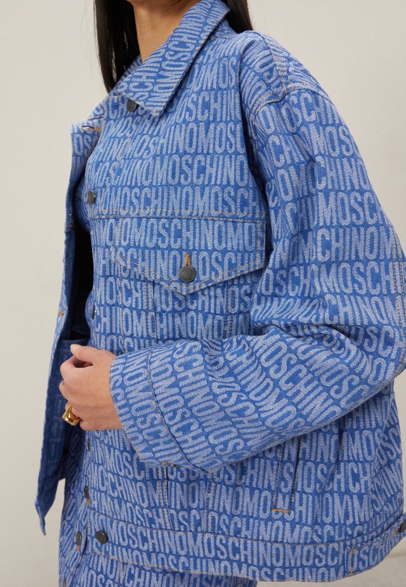 Primer plano de una persona que lleva una chaqueta y una falda de mezclilla azul cubiertas con un patrón de texto repetido "MOSCHINO", con la mano tocando la parte delantera de la chaqueta.