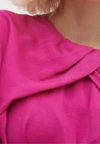 Fuksia-bluusi pehmeästä, virtaavasta kankaasta ja laskostetusta kaula-aukosta. Pinnassa on sileä rakenne, jossa on hienovaraisia ryppyjä ja taitoksia.