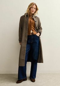 Cappotto lungo a quadri in marrone e crema, indossato sopra un cardigan marrone, con jeans a gamba larga blu scuro e scarpe marroni, in piedi contro uno sfondo chiaro.