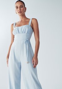 Jasnoniebieski kombinezon z miękkiej tkaniny z kwadratowym dekoltem, marszczoną górą, szerokimi nogawkami i paskiem w talii z złotymi akcentami.