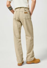 Beige denim pants feature a straight leg design, two back pockets, and a branded leather patch. Worn with tan boots and a neutral shirt.