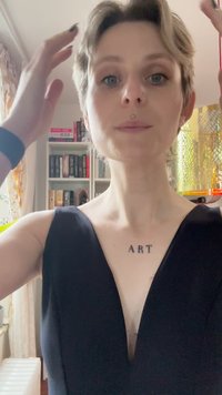 Fekete V nyakú ruha, testhezálló felső résszel. Rövid haj, látható tetoválások a nyakon. A háttérben könyvespolcok és egy dekoratív lámpa található.