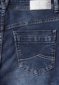 Jean en denim bleu marine avec une poche arrière présentant une forme pointue distinctive et des coutures blanches contrastées. Texture lisse, design minimal.
