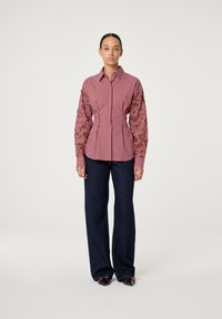 Gestreept rood-wit shirt met gestructureerde bruine borduursels op de mouwen, gecombineerd met donkerblauwe wijde jeans en bruine schoenen.