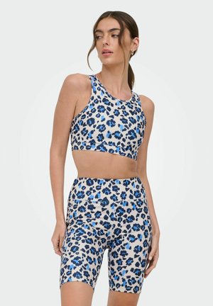 Kobieta ubrana w niebiesko-czarny top na ramiączkach w panterkę i dopasowane, wysokie szorty rowerowe, stojąca na jasnym, jednolitym tle.