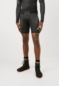 Shorts compressivi neri realizzati in un materiale liscio ed elastico, con piping a contrasto e un logo sulla coscia. Abbinati a scarpe sportive nere.