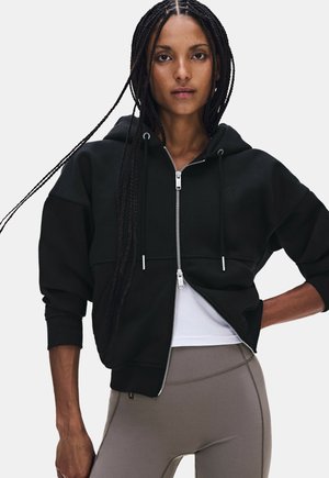 Femme avec des cheveux tressés portant un sweat à capuche noir zippé par-dessus un top blanc et des leggings gris, debout avec une main sur la hanche sur fond blanc.
