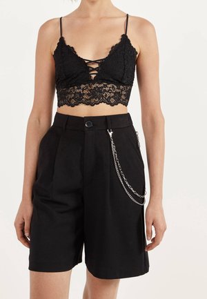 Czarny koronkowy biustonosz o krzyżowym wzorze i falbaniastym wykończeniu, zestawiony z czarnymi szortami o wysokim stanie z akcentem w postaci łańcuszka.