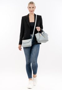 Tan leren handtas en een lichtblauwe crossbodytas, beide met gouden accenten, vastgehouden door een vrouw in een zwarte blazer en skinny jeans.