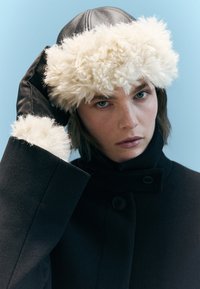 Czarny wełniany płaszcz z wysokim kołnierzem, z detalem w postaci guzik�ów; skórzane rękawiczki; puszysty biały kapelusz z futra z teksturowanym brzegiem; neutralne tło.