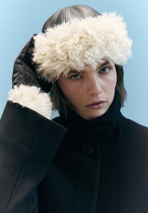 Zwarte wollen jas met hoge kraag, voorzien van knoopdetails; leren handschoenen; pluizige witte bontmuts met gestructureerde rand; neutrale achtergrond.