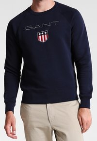 Felpa blu navy con collo a giro, caratterizzata da un logo "GANT" in rilievo e un emblema a scudo con stelle. Realizzata in tessuto testurizzato, dal design aderente.