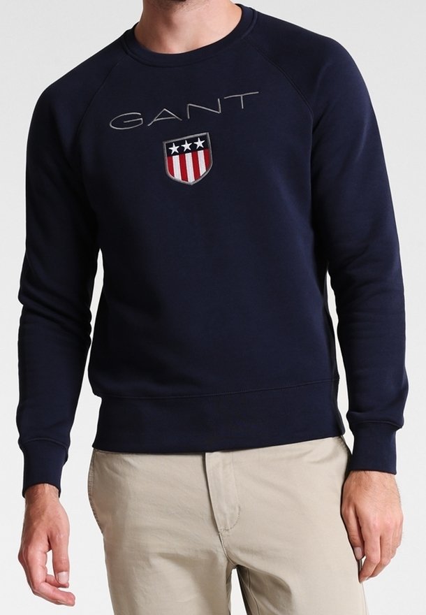 Felpa blu navy con collo a giro, caratterizzata da un logo "GANT" in rilievo e un emblema a scudo con stelle. Realizzata in tessuto testurizzato, dal design aderente.