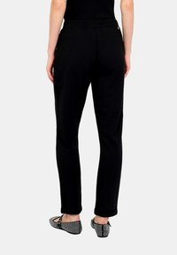 Persona che indossa pantaloni neri a sigaretta e scarpe basse con motivo bianco e nero, in piedi contro uno sfondo semplice, vista posteriore.