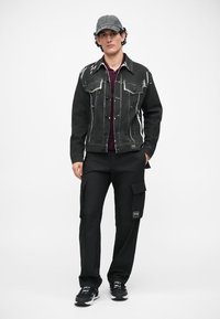 Μαύρο τζάκετ από τζιν με λευκές ραφές, συνδυασμένο με μαύρο cargo παντελόνι με πλαϊνές τσέπες, φορεμένο με γκρι καπέλο και αθλητικά παπούτσια.