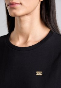 Black t-shirt with a round neckline, featuring a small gold metal logo badge positioned on the left side near the neckline.