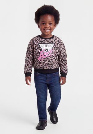 Bambino sorridente che cammina, indossa una felpa Guess con stampa leopardata e la scritta "Love", jeans blu e scarpe slip-on nere su sfondo bianco.