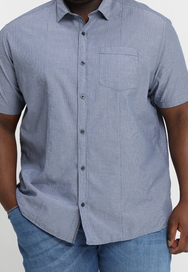 Homme portant une chemise bleu clair à manches courtes boutonnée avec une poche poitrine et un jean bleu, les mains détendues le long du corps.