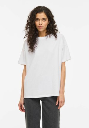 Kvinne med krøllete mørkt hår iført en løs hvit kortermet skjorte og svarte bukser, stående foran en ensfarget hvit bakgrunn.