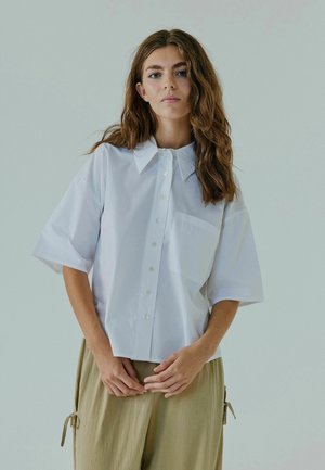 Hvid kortærmet skjorte med spids krave, frontknapper og en brystlomme, parret med lyse beige løstsiddende bukser.