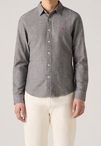 Camicia grigia con bottoni, realizzata in tessuto testurizzato, dotata di colletto classico e logo Levi's sul petto; abbinata a pantaloni chiari.