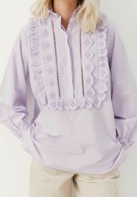 Blusa di un leggero lavanda con rifiniture in pizzo, ritagli decorativi e colletto alto. Le maniche lunghe presentano dettagli arricciati ai polsini.