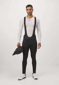 Schwarze Bib-Radsporthosen mit Trägern und gepolstertem Sitz. Getragen mit einem weißen Langarm-Basisshirt und einem schwarzen Helm.