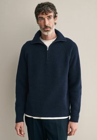 Pulover cu guler înalt, de culoare bleumarin, cu textură canelată, prevăzut cu fermoar la gât și mâneci lungi. Purtați peste o cămașă albă, pe un fundal neutru.