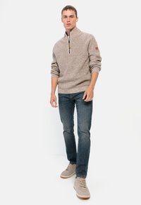 Beiger Strickpullover mit hohem Kragen und Reißverschluss, kombiniert mit dunkelblauen Jeans und hellgrauen Sneakern, mit einem strukturierten Muster.