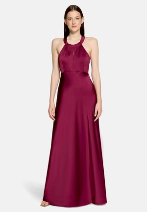 Kobieta ubrana w bezrękawnikową, sięgającą podłogi, bordową sukienkę z dekoltem halter, stojąca na białym tle.