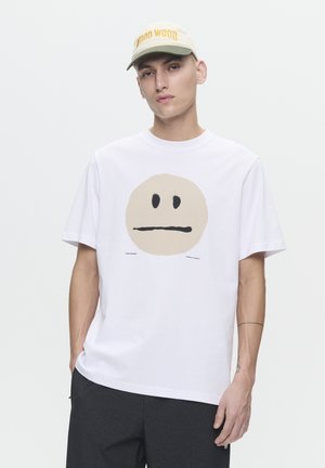 White cotton t-shirt featuring a large, tan circular face graphic with a neutral expression. Paired with a beige and green cap.
