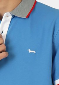 Polo shirt azzurro con logo di un bassotto bianco. Presenta un colletto in rete grigio con un accento rosso. Materiale in cotone. Maniche corte.