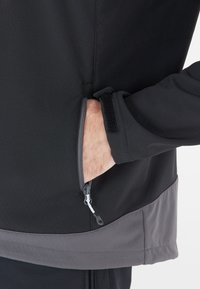 Schwarze Softshell-Jacke mit grauen Akzenten; verfügt über eine Reißverschlusstasche und einen Klettverschluss am Ärmel. Glatte Textur und körperbetontes Design.