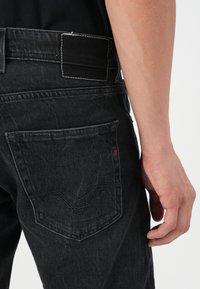 Jeans de denim negro con un diseño ajustado, que presentan un bolsillo trasero con detalles bordados y costuras blancas en contraste.