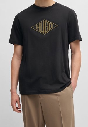 Mand iført sort T-shirt med korte ærmer med brunt "HUGO"-logo i diamantform og beige bukser, hånd i lommen, foran en ensfarvet baggrund.