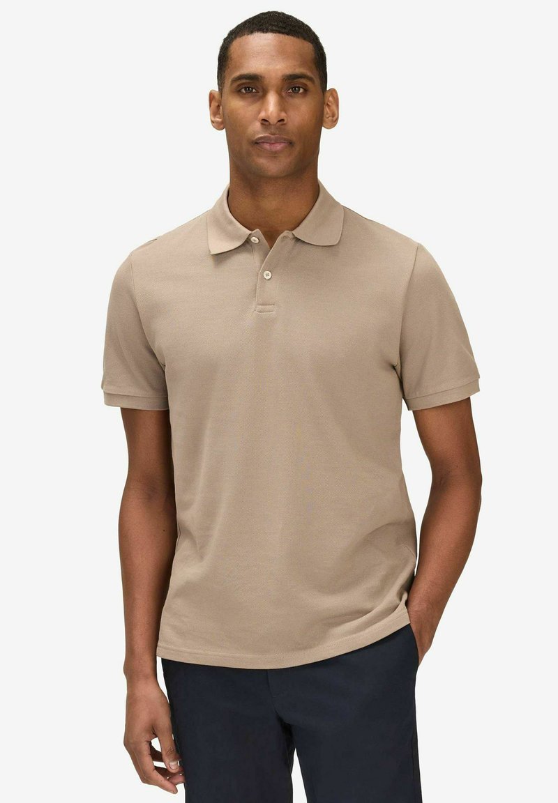Homme portant un polo beige à manches courtes et un pantalon foncé, debout avec une main dans la poche devant un fond clair uni.
