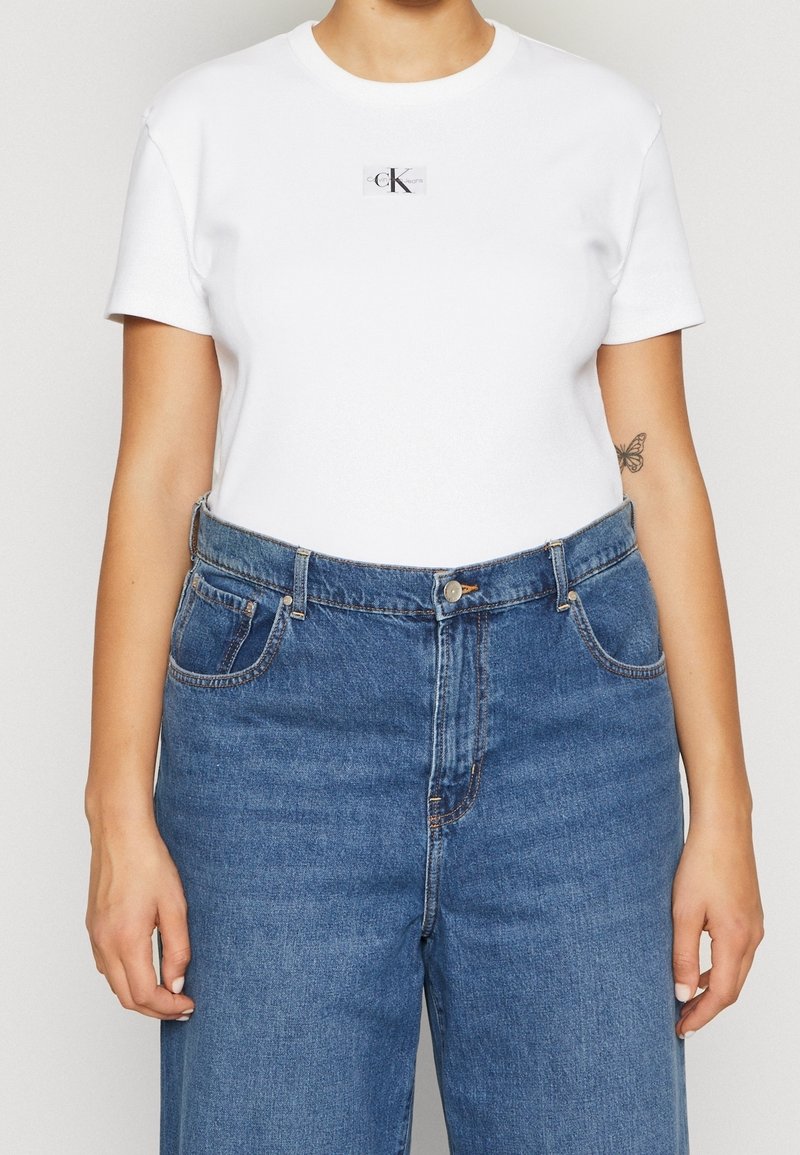 T-shirt bianco in cotone con maniche corte, con un piccolo logo nero. Abbinato a jeans blu in denim a vita alta, con una vestibilità rilassata.