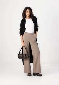 Svart lang cardigan, hvit skjorte, beige vide bukser, brun håndveske og mørke sko. Krøllete hår; nøytral bakgrunn.