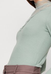 Rippierter, hellgrüner Rollkragenpullover mit einer strukturierten Spitze-Kragen, kombiniert mit hoch taillierten, hellbraunen Hosen, die einen eleganten Reißverschluss haben.