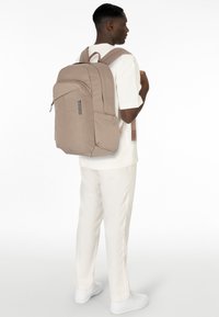 Mochila beige con un diseño estilizado, material de tela, compartimentos con cremallera y un logo en la parte frontal. Correas ajustables y acolchadas.
