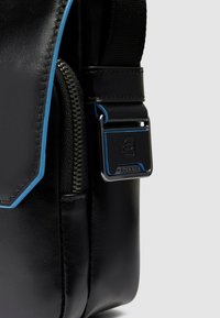 Borsa in pelle nera con rifiniture blu, dotata di chiusura a cerniera, dettagli metallici e superficie texturizzata. Tracolla regolabile inclusa.