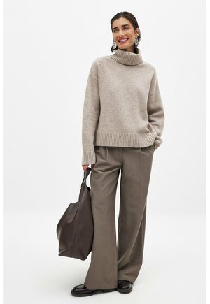 Beige Rollkragenpullover mit geripptem Saum, kombiniert mit lockeren braunen Hosen. Das Modell hält eine dunkelbraune Ledertasche. Glatte Texturen, entspannter Schnitt.