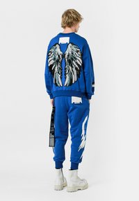 Sudadera azul con diseño de alas de lentejuelas en la parte posterior, combinada con pantalones de chándal azules a juego que cuentan con acentos blancos y una etiqueta decorativa.