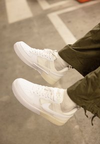 Vita lädersneakers med perforerade tår och transparenta accenter, kombinerade med ljusgrå strumpor och olivgröna byxor.