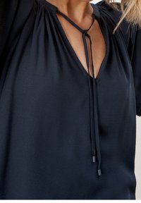 Haut texturé bleu marine avec décolleté en V et fermeture à nouer, présentant des détails froncés près de l'encolure et des manches courtes.