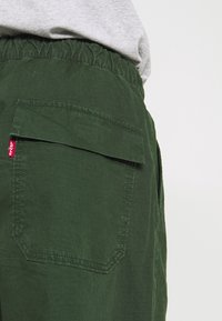 Pantaloni Levi's verdi con cintura elastica e tasca a patta, indossati con una camicia grigia chiara visibile in vita.