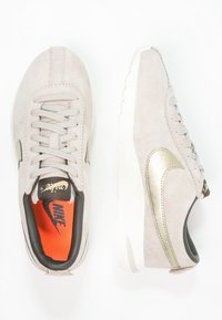 Vaaleanharmaat mokkatossut, joissa on kultaiset yksityiskohdat, pyöreä kärki ja kumipohja; sisältää mustan ja oranssin sisustan. Nyöritys suljenta, jossa on teksturoitu pinta.